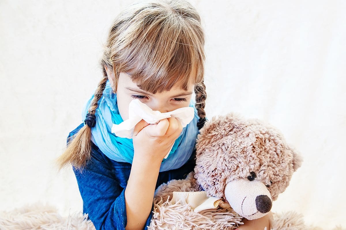 A little girl is sneezing into a tissue paper while holding a toy dog in her other hand.